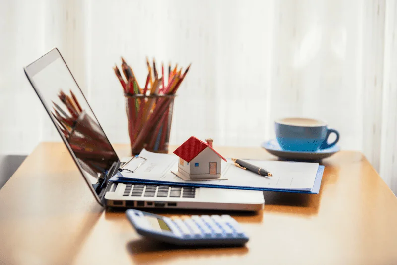 A model home sits on top of a contract and laptop on a desk with a calculator and cup of pencils.