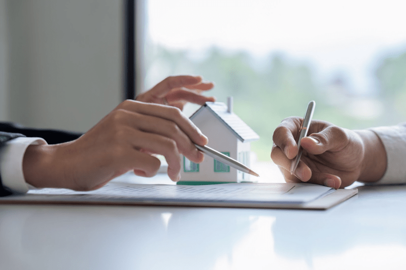 Two people with pens in hand review a contract sitting next to a small model home