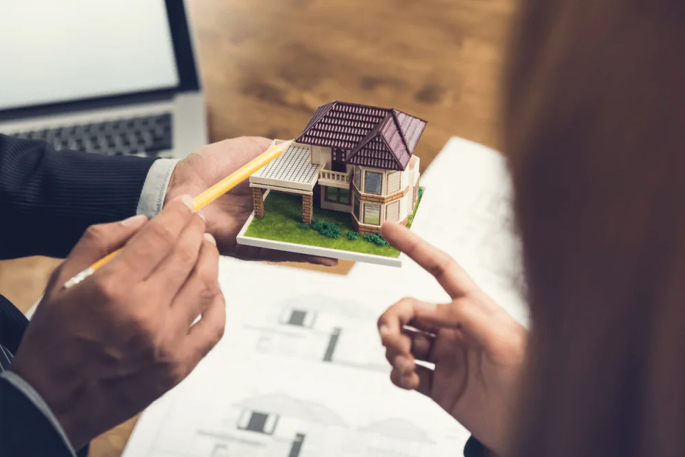 A person gestures towards a small model home using a pencil.