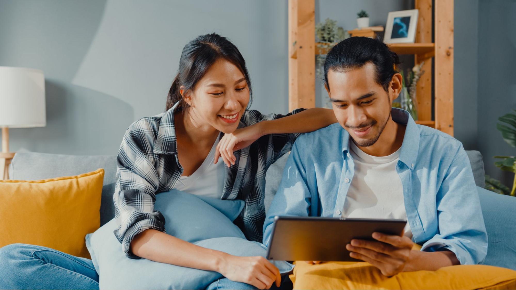 A smiling couple on the couch in their home looking at a tablet. 