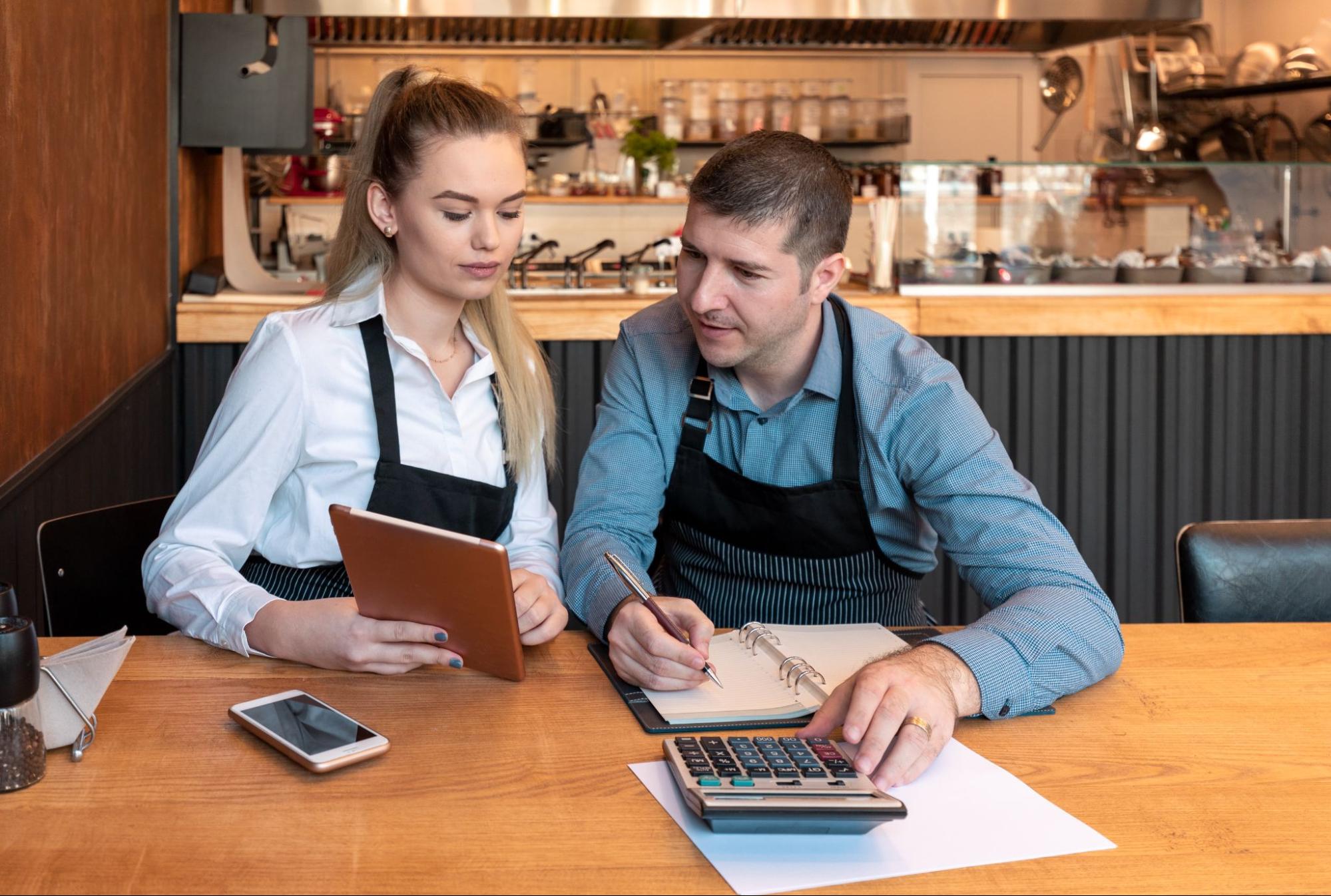 A male and a female small business owner sit in their cafe and consult a tablet while organizing their finances.