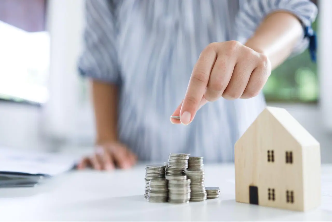 A person stacking coins next to a small wooden model of a home.