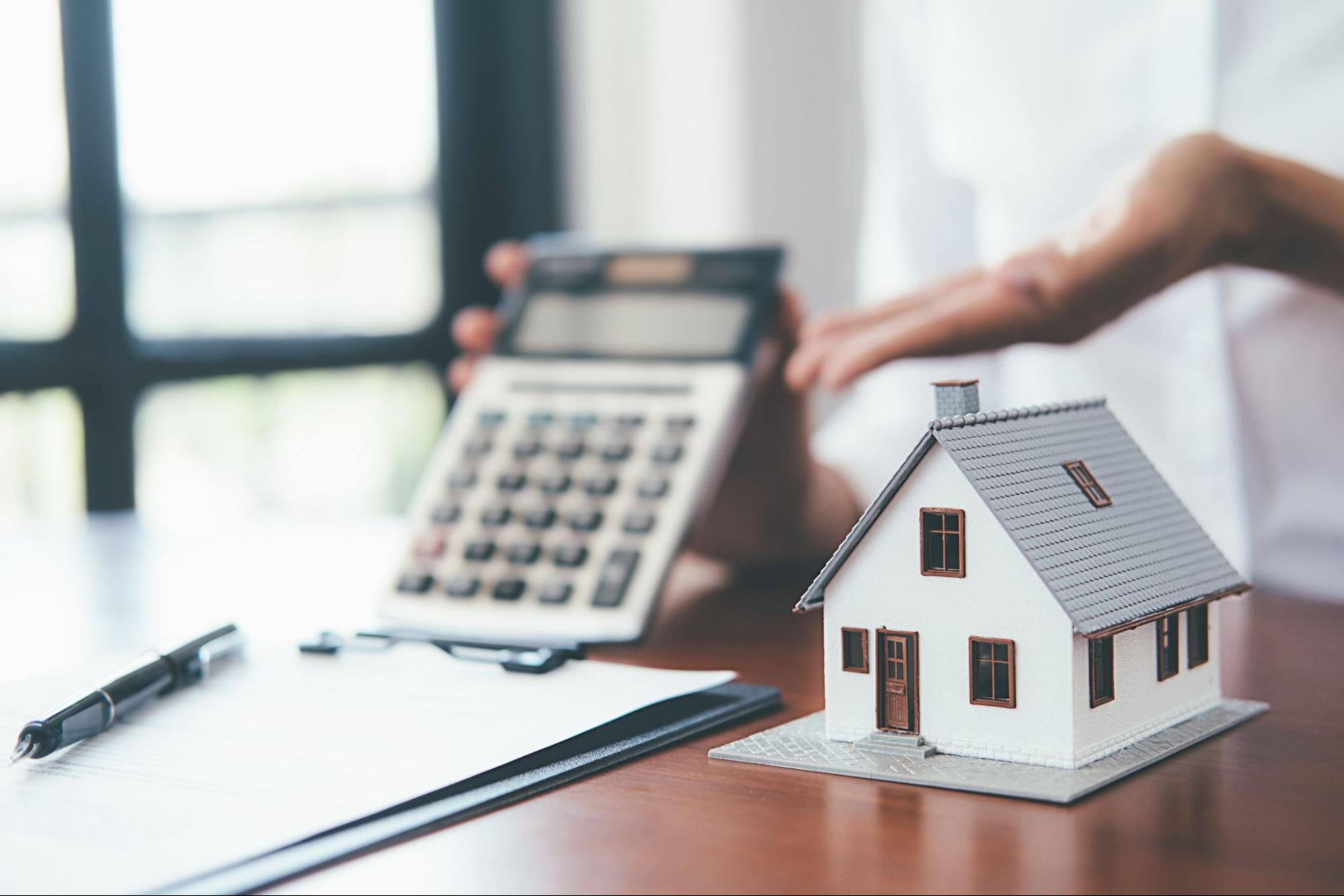 A person holds up a calculator next to a small model home.