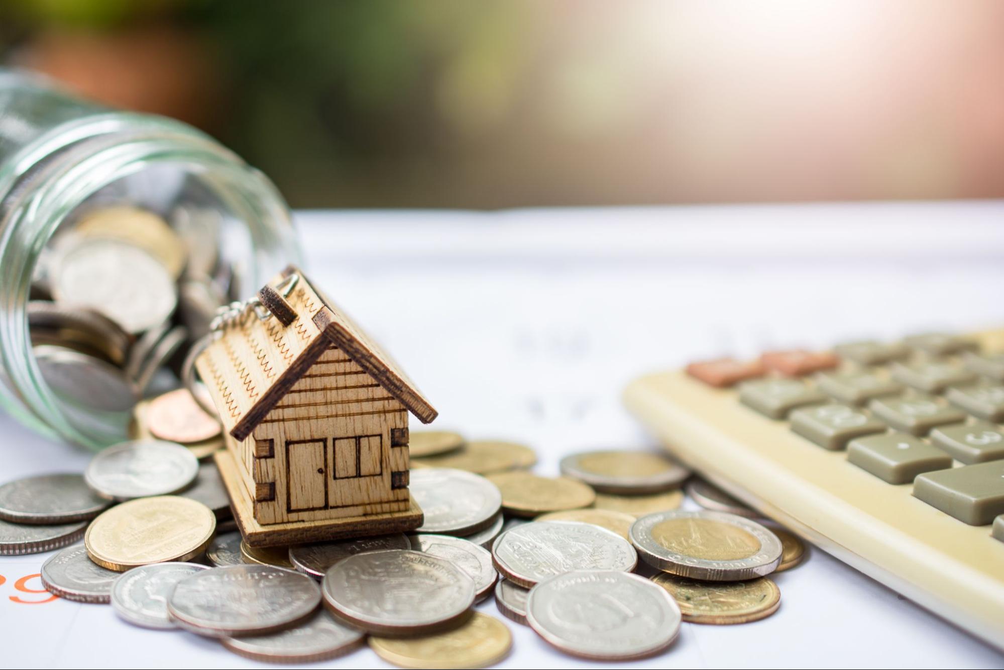 A wooden model home on top of a pile of coins next to a calculator.