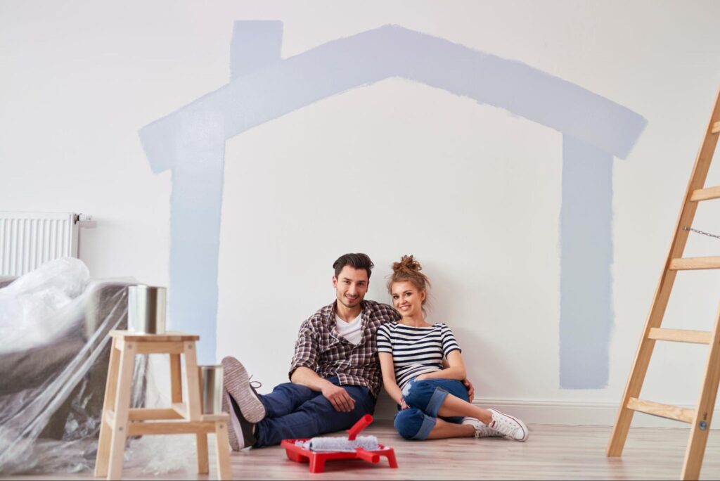 A young couple sits in their home mid-remodel beneath the painted outline of a house on the wall.