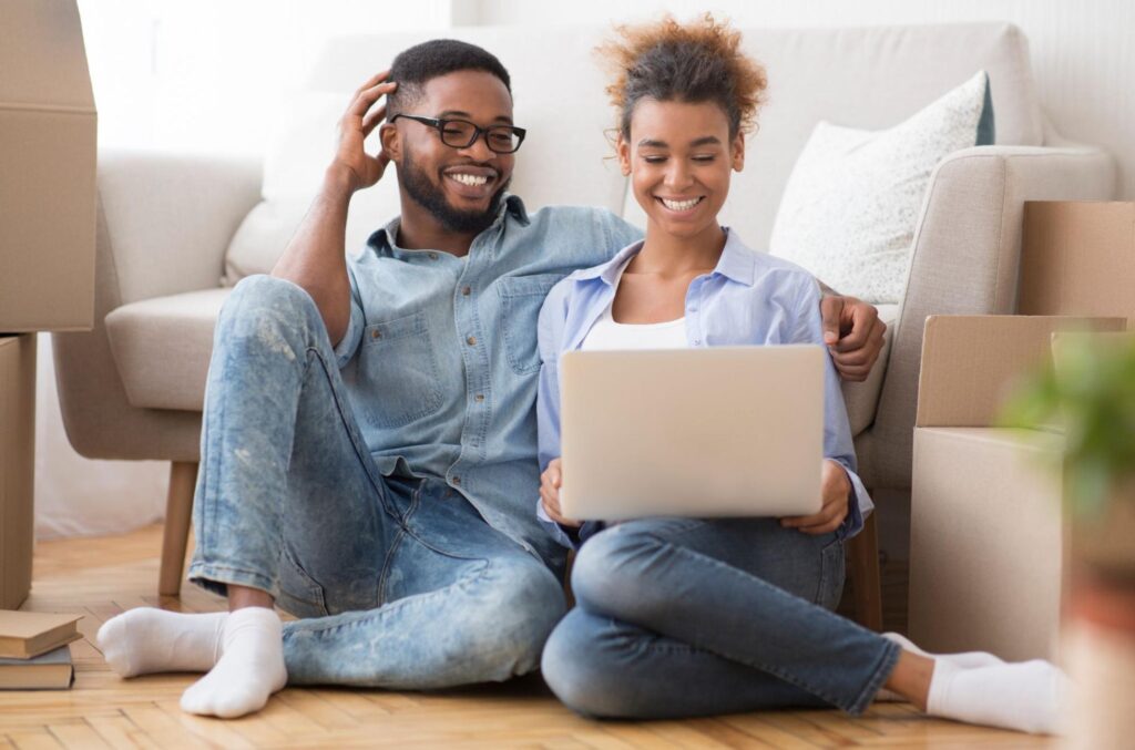 A young smiling couple sit on their living room floor and look at their laptop.