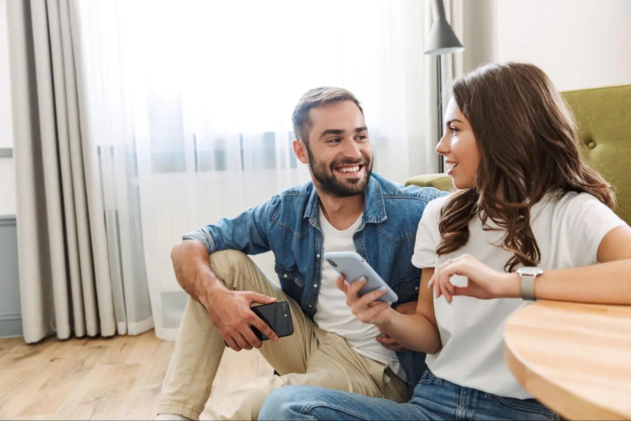 A smiling young couple sitting in the living room on their phones.