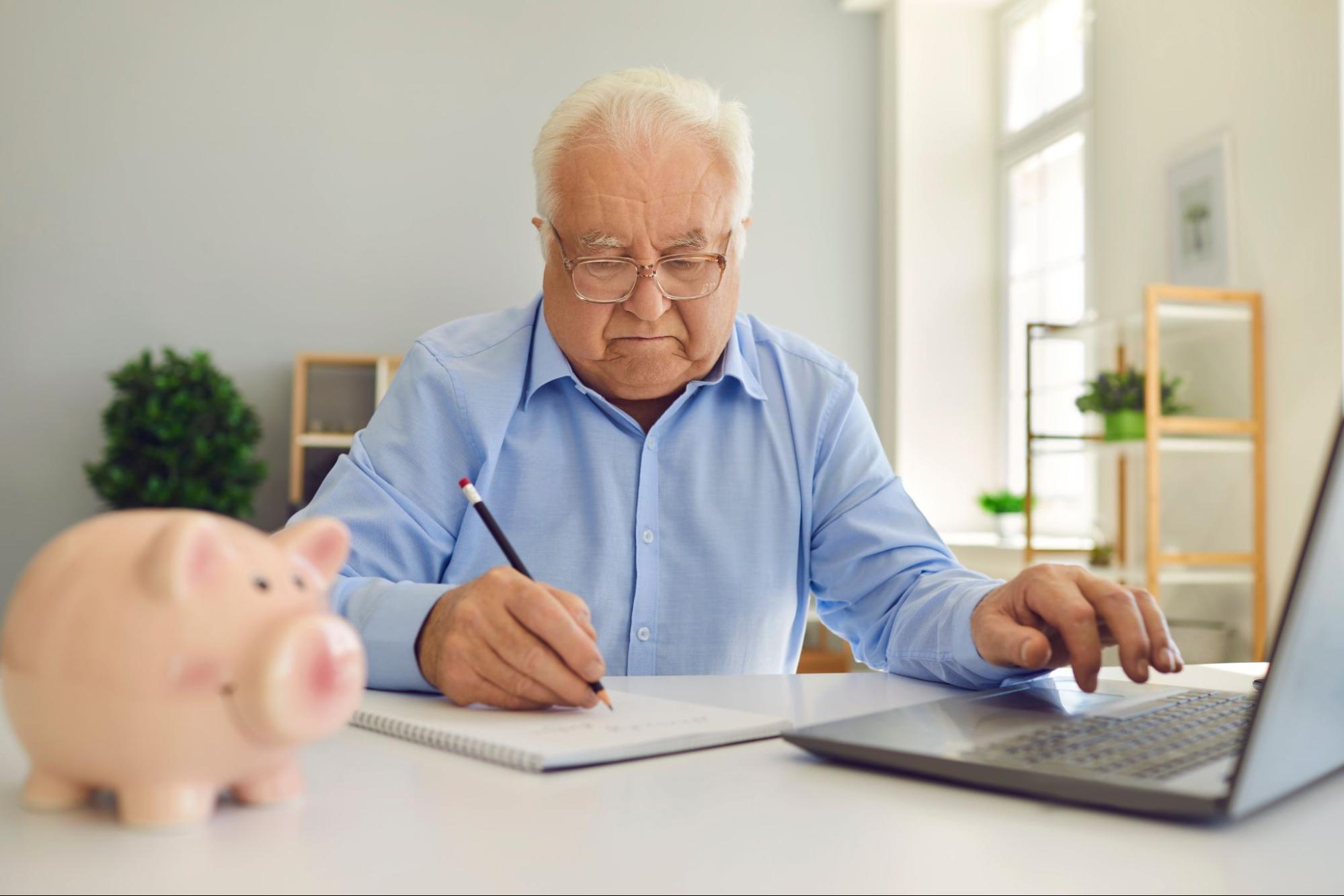 An elderly man next to a piggy bank and laptop writing in a notebook.