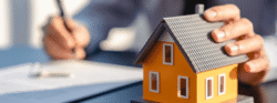 A person fills out a mortgage document and holds a small orange model home.