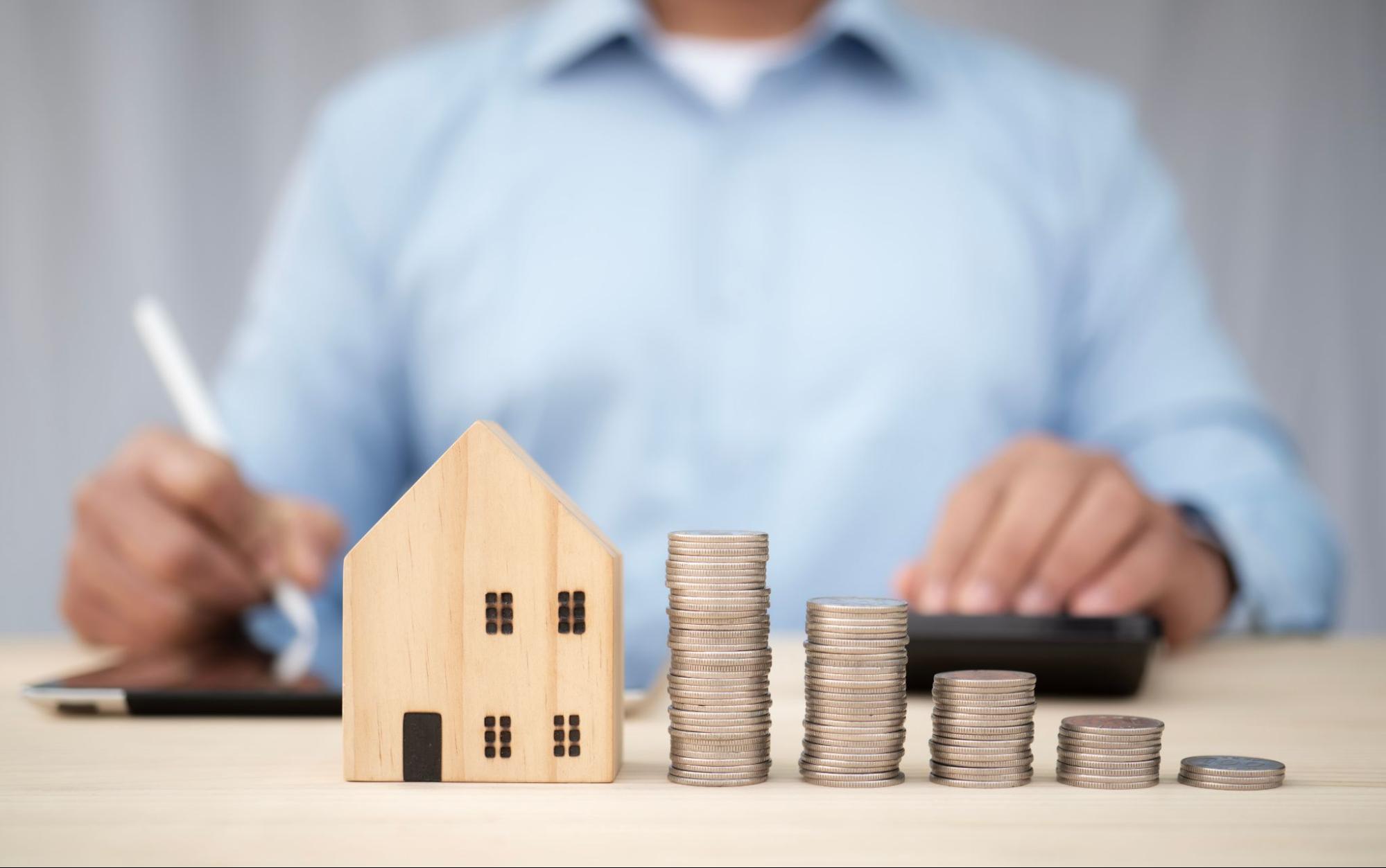 A person writes on a tablet while sitting behind a model home next to stacks of coins.