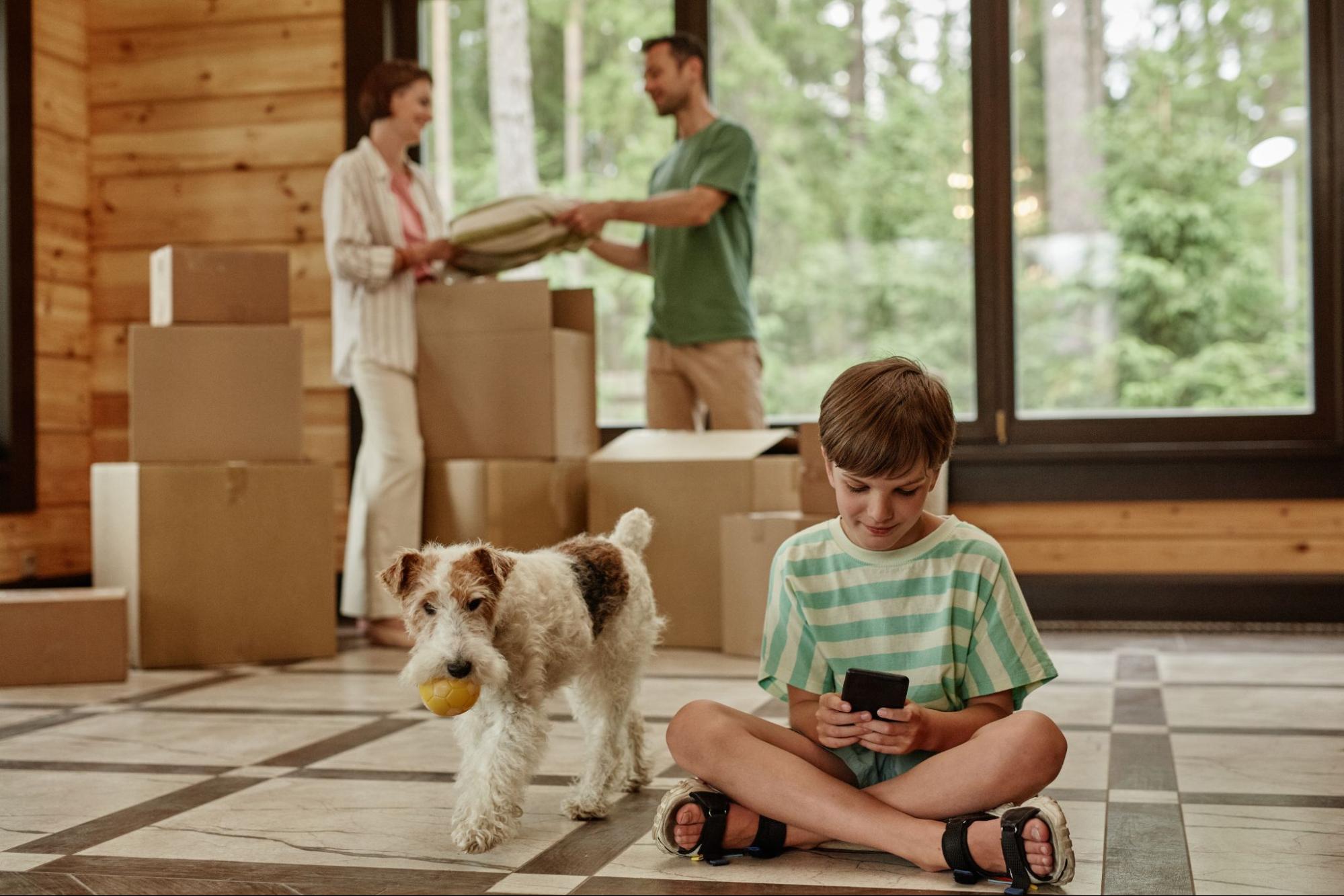 A family unpacks in their new home while the dog roams around and their son plays on his phone.