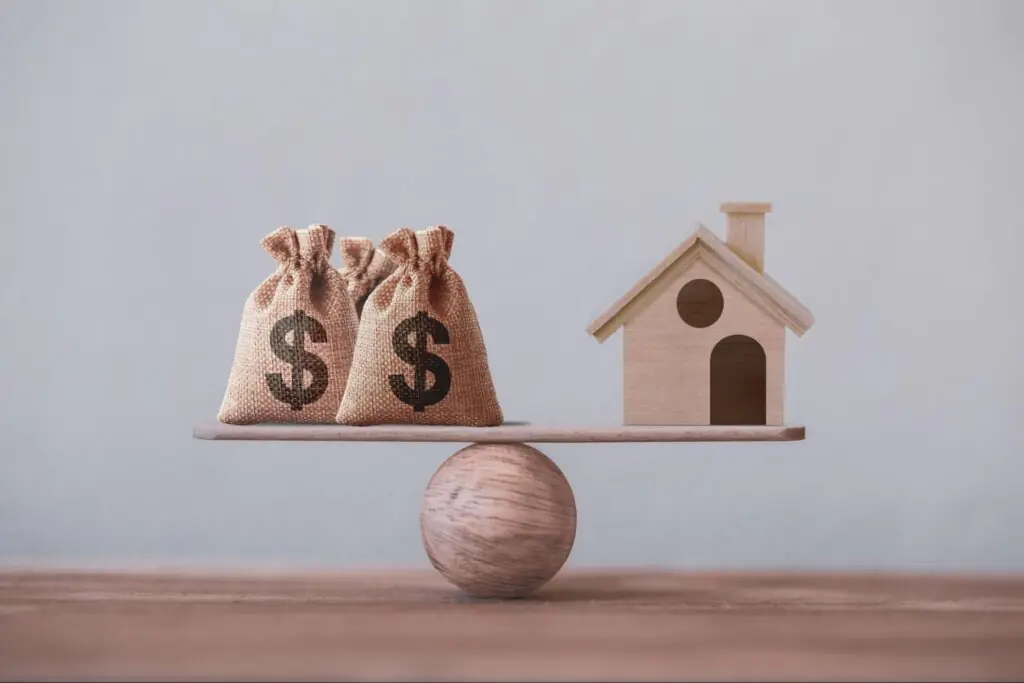 Bags of money and a small model home on opposite sides of a balance scale.