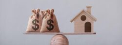 Bags of money and a small model home on opposite sides of a balance scale.