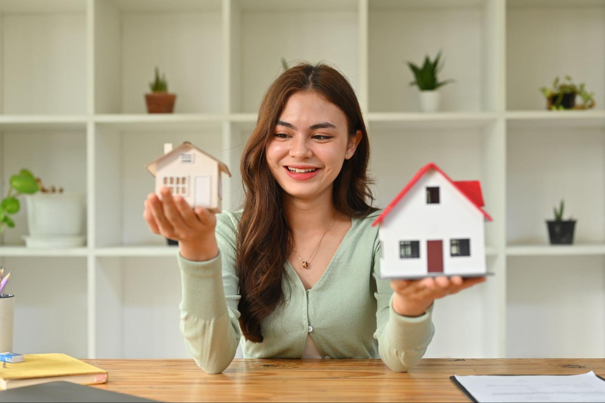 A young woman at a desk holds a small model home in each hand.