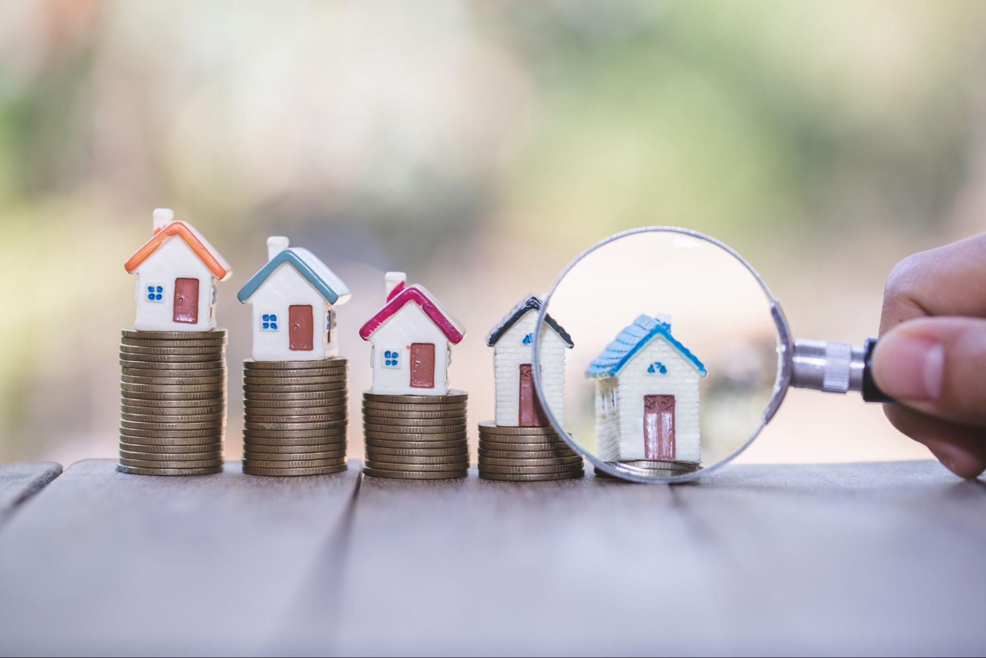A magnifying glass examining model homes that are placed on stacks of coins.