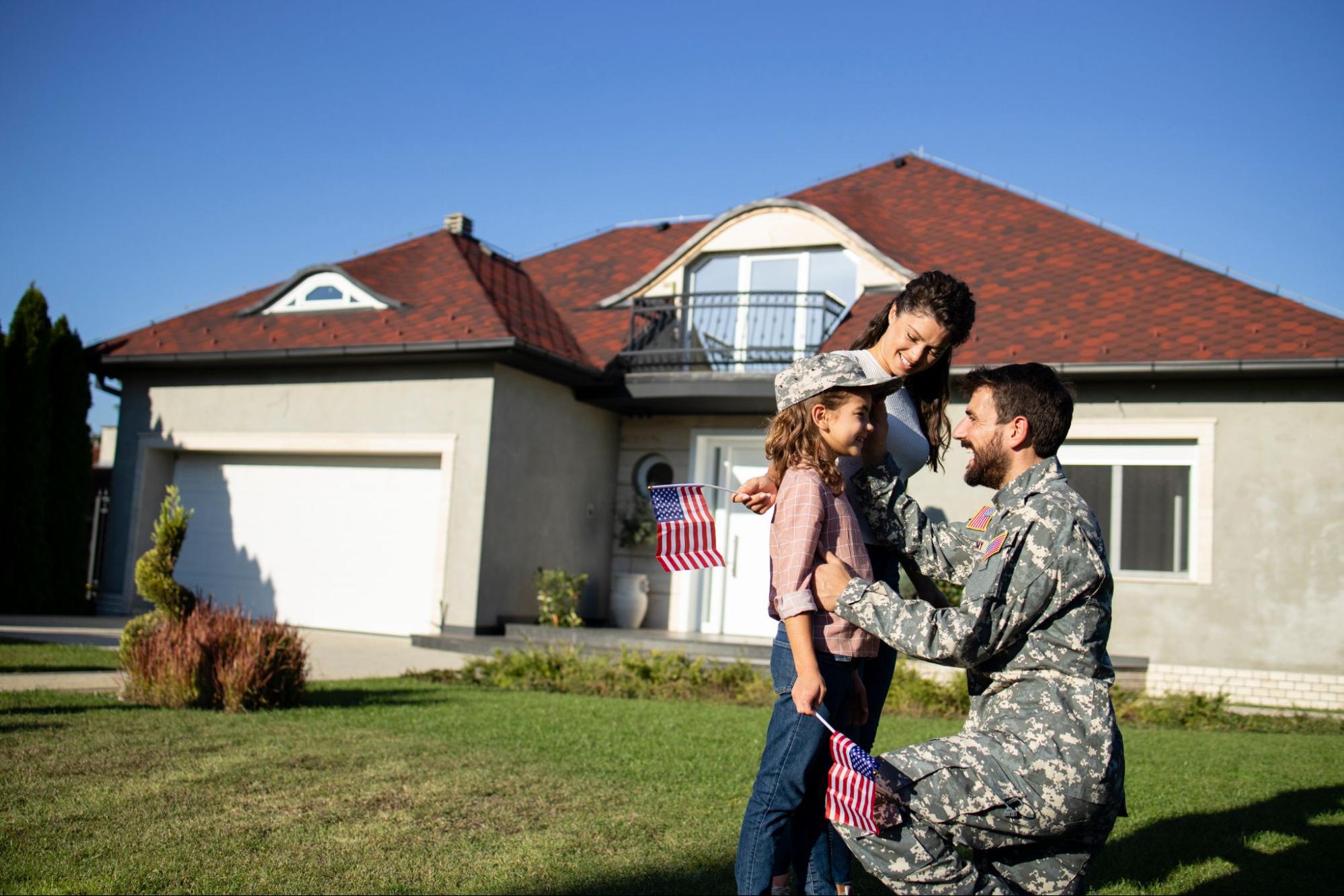 A service member in uniform embracing his daughter and wife on the lawn outside their home. 