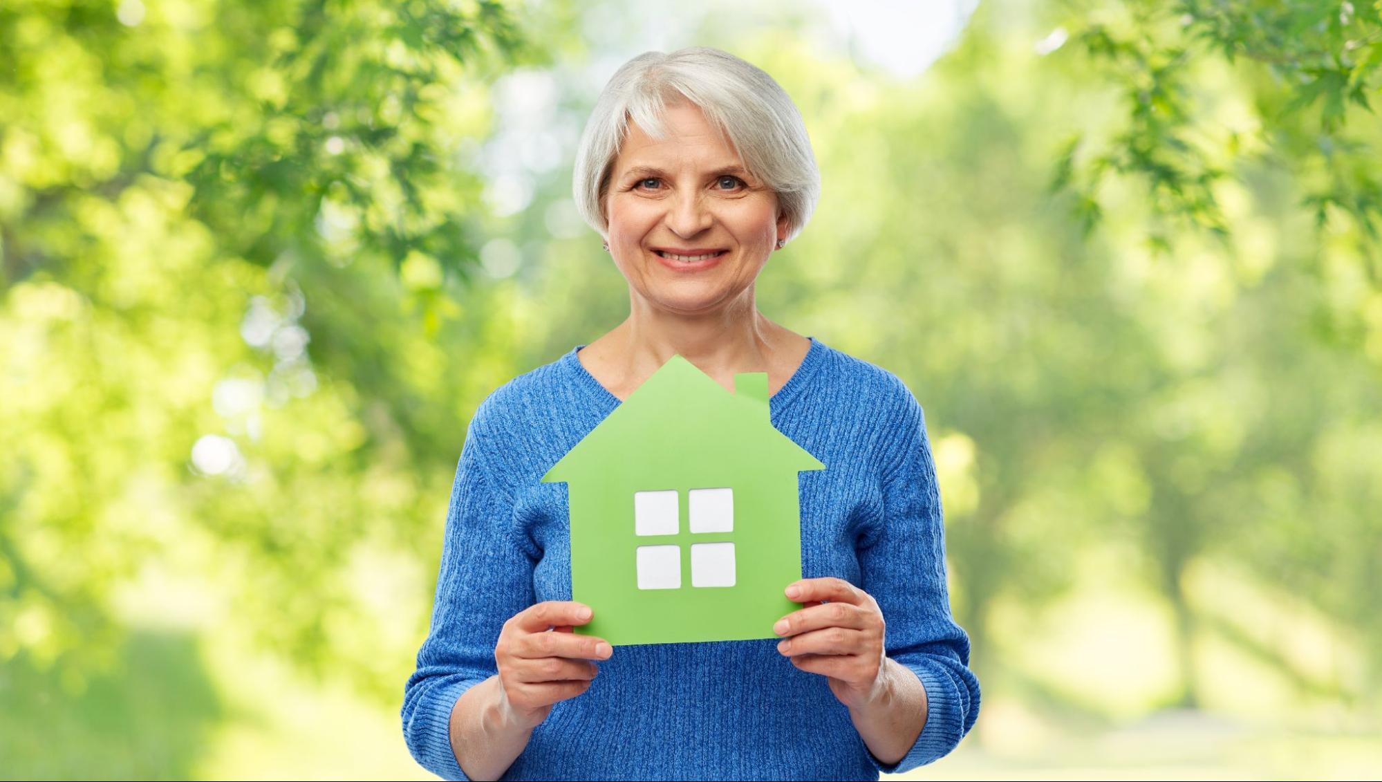An elderly woman in a blue sweater holds up a green cutout in the shape of a home.Â