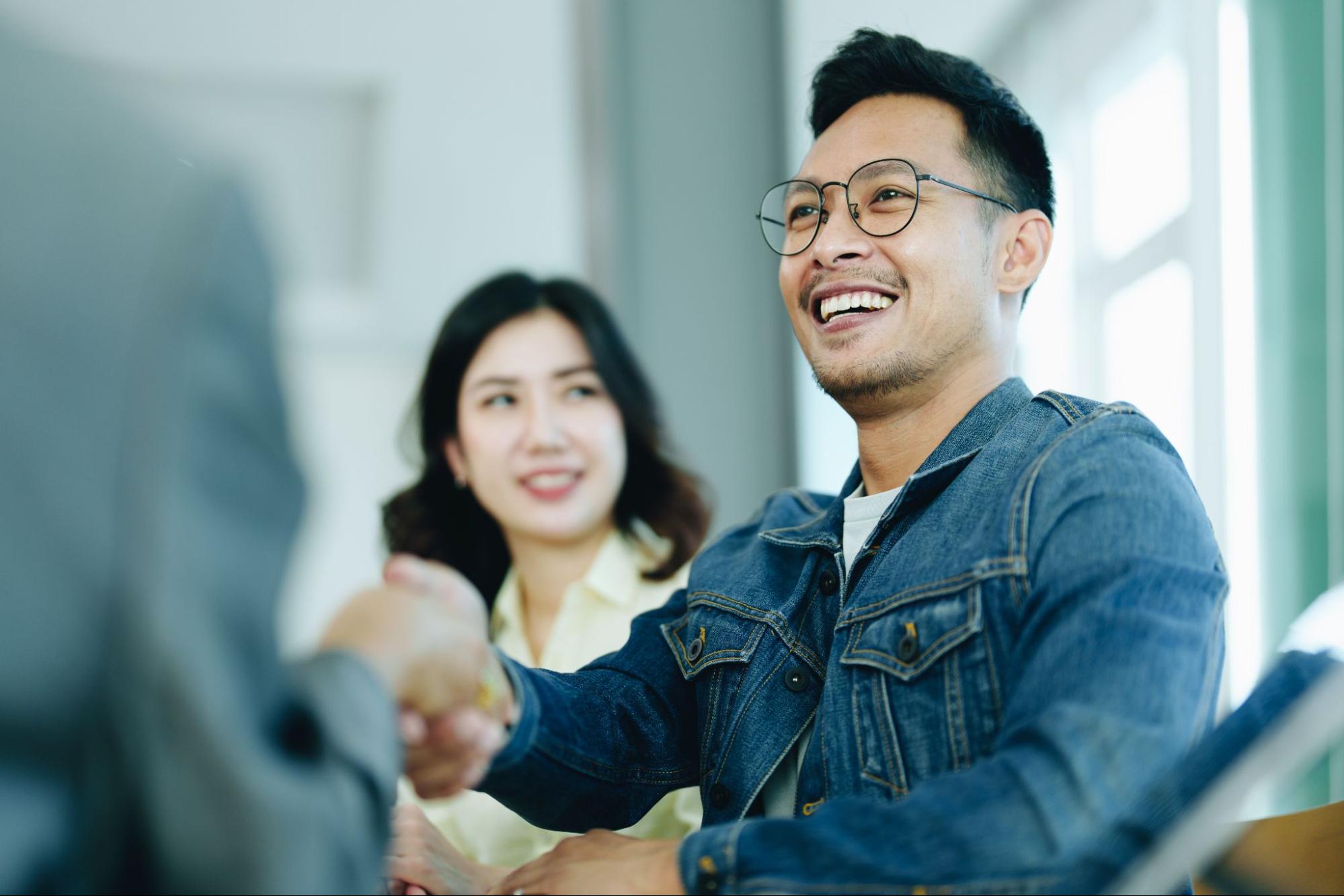 A young Asian man wearing glasses shakes hands with his mortgage lender. 
