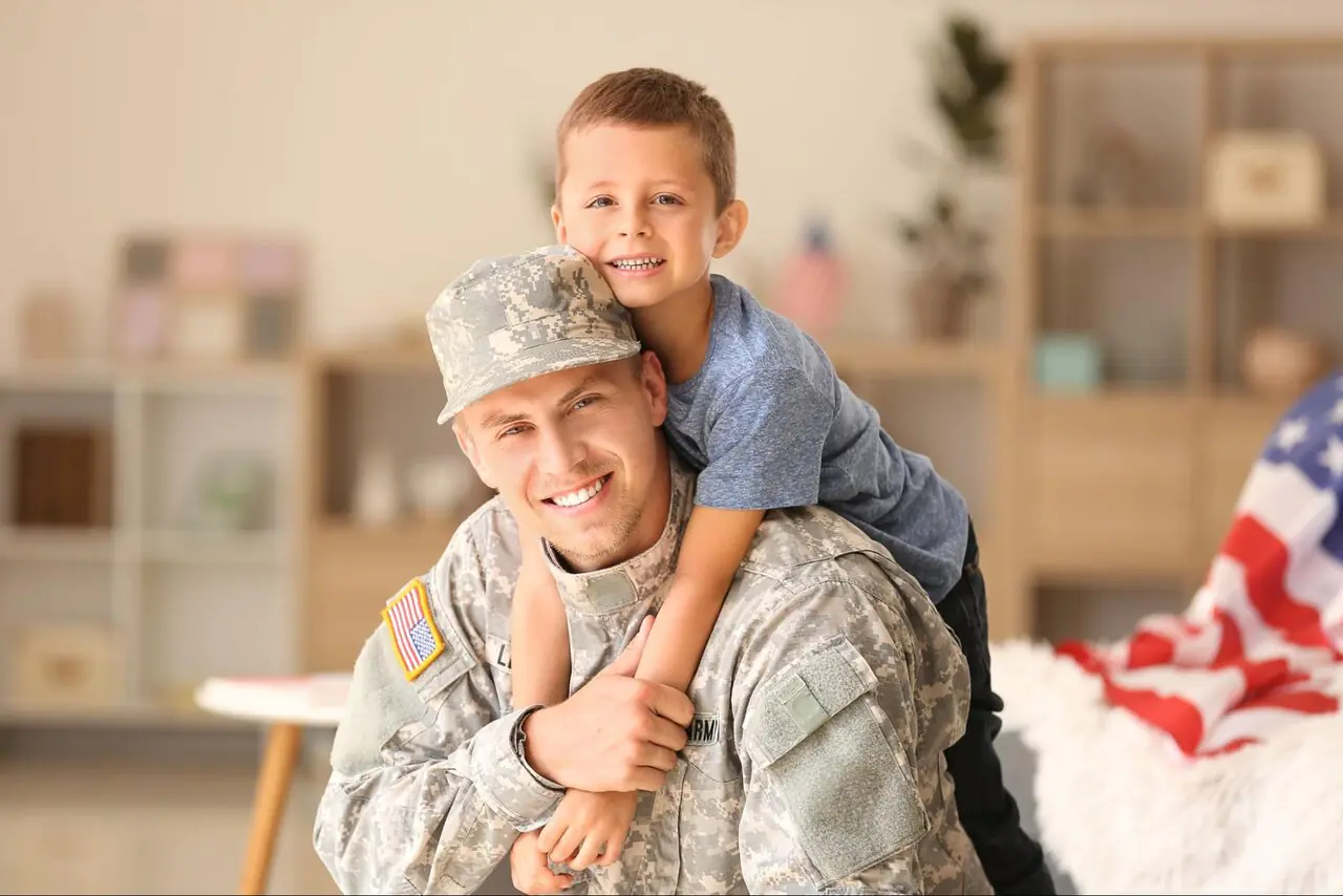 A male service member smiling with his son’s arms wrapped around his shoulders.