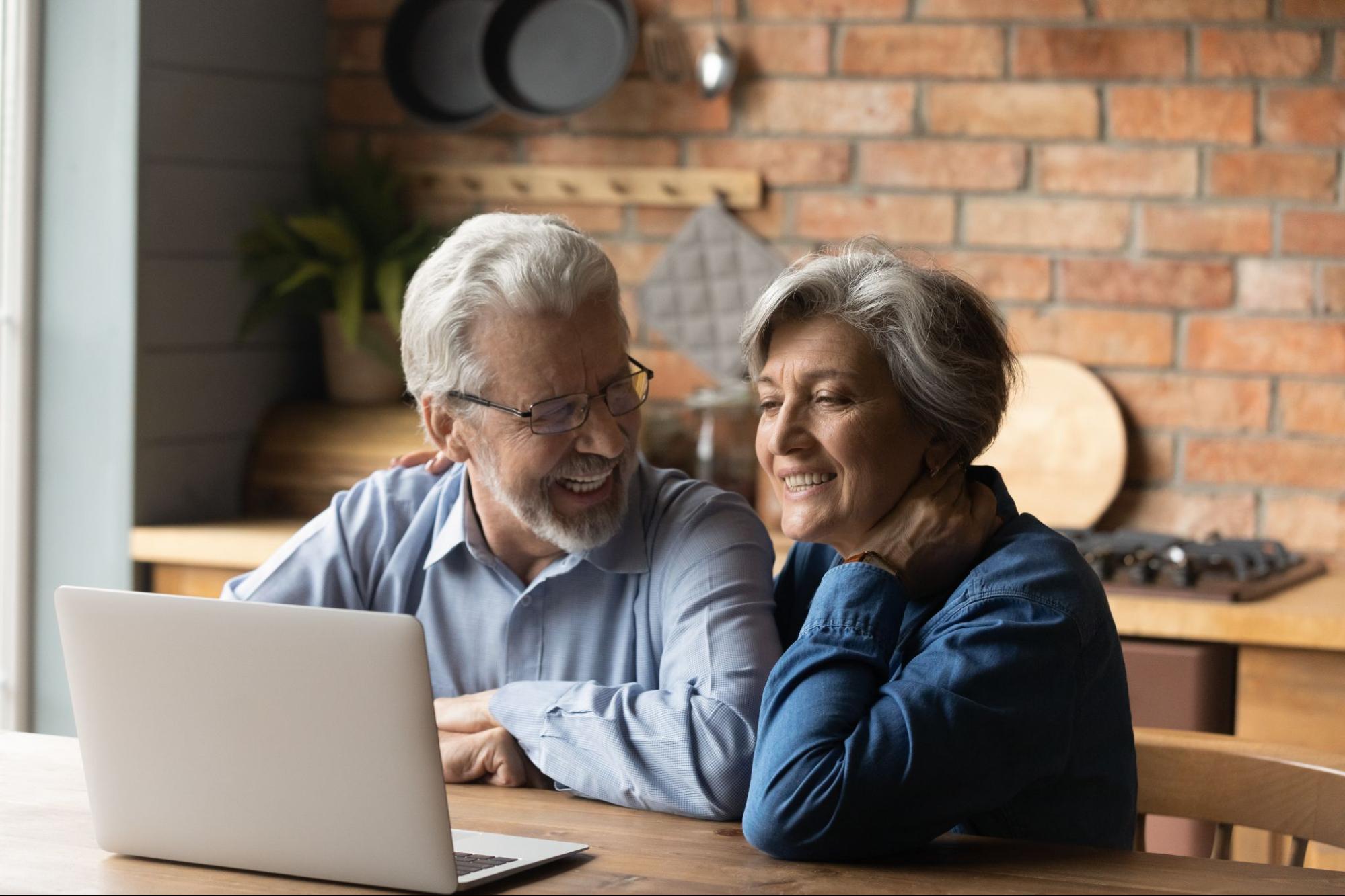 A smiling older couple sitting in their kitchen and looking at their laptop. 