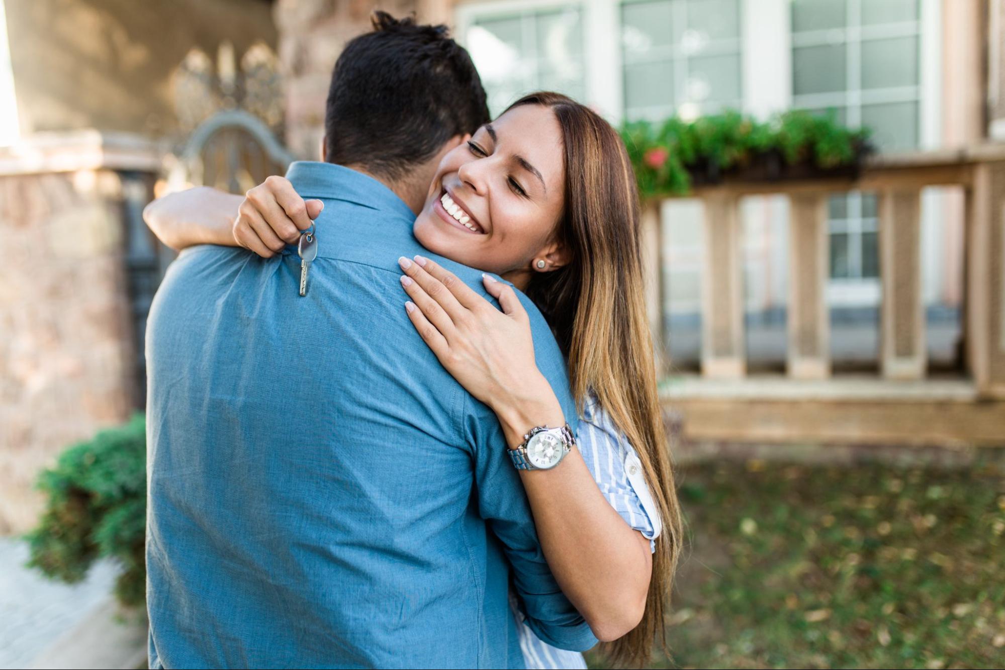 A smiling man and woman hug in front of the home they just purchased. 