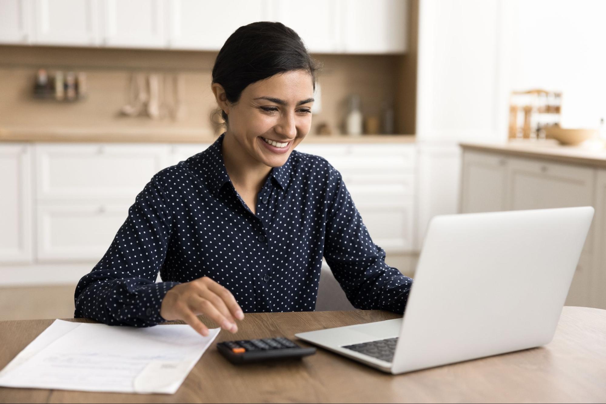 A smiling woman looking at her laptop and using a calculator. 