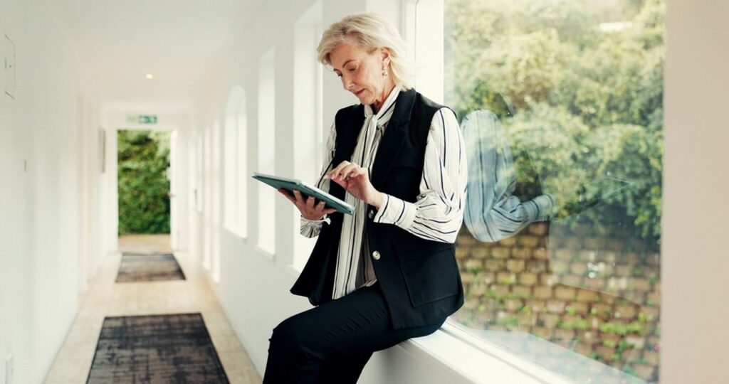 Older businesswoman using a tablet while sitting beside a large window in a bright modern office.